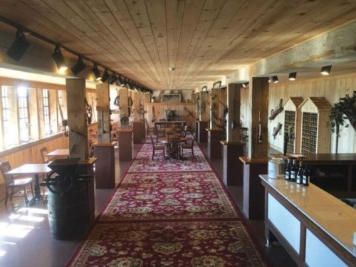 A rustic interior of a winery with wooden beams, tables, and a patterned rug, featuring wine bottles on display.