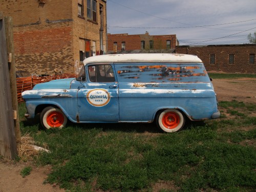 A vintage blue delivery van with orange wheels, parked in a grassy area near old brick buildings.