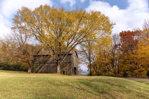 A rustic barn surrounded by vibrant autumn trees and a grassy hillside under a partly cloudy sky.
