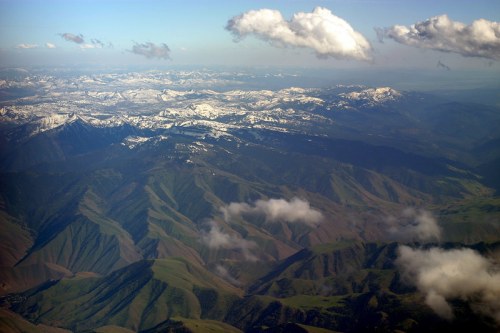 Aerial view of snow-capped mountains and rolling green hills under a clear blue sky with scattered clouds.