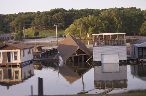 Houses partially submerged in floodwaters, with one structure leaning precariously, surrounded by trees and power lines.