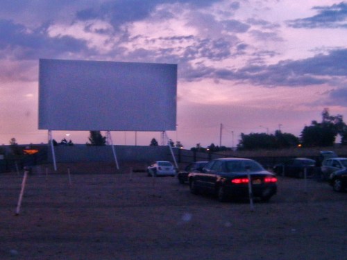 A drive-in movie screen at dusk, with parked cars in the foreground and a colorful sky in the background.