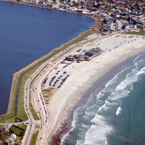 Aerial view of a beach with waves, a parking lot, and a road alongside a coastal town.