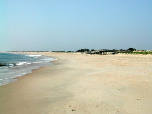 A serene beach scene with gentle waves, soft sand, and a distant boardwalk under a clear blue sky.