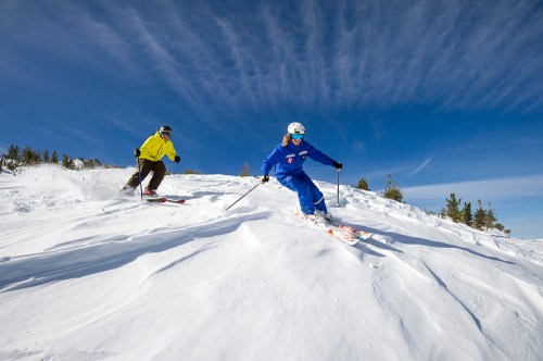 Two skiers navigate a snowy slope under a clear blue sky, with trees in the background and wispy clouds above.