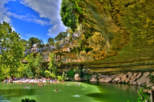 A scenic view of a natural swimming area with people enjoying the water, surrounded by lush greenery and rocky cliffs.