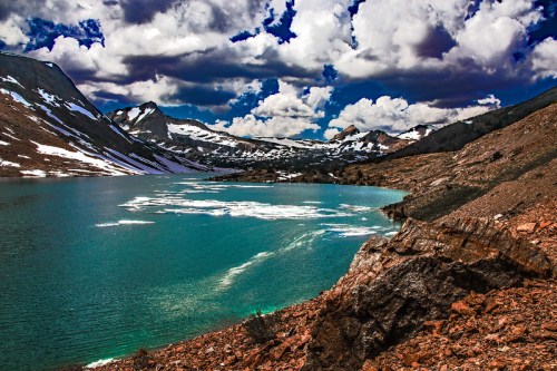 A serene mountain lake surrounded by rocky terrain and snow-capped peaks under a dramatic sky with fluffy clouds.