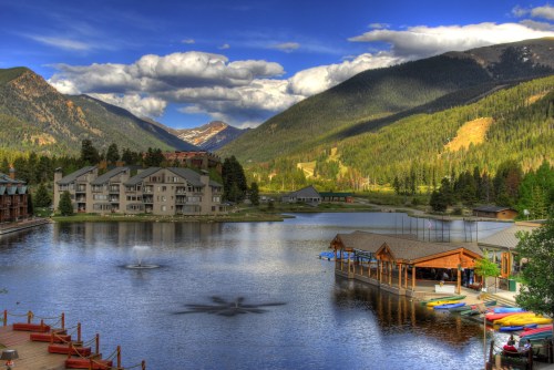 Scenic view of a lake surrounded by mountains, with a resort, kayaks, and a fountain under a blue sky with clouds.
