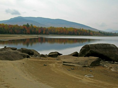 A serene lake surrounded by autumn foliage and mountains, with rocks in the foreground and calm water reflecting the scenery.