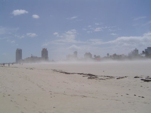 A sandy beach with a misty skyline in the background, featuring tall buildings and a clear blue sky.