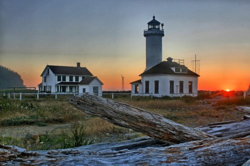 A lighthouse and a nearby house at sunset, with a log in the foreground and a serene landscape in the background.