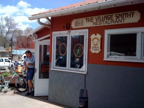 A man stands outside The Village Smithy Restaurant, with a bike nearby and a sign indicating it's open.