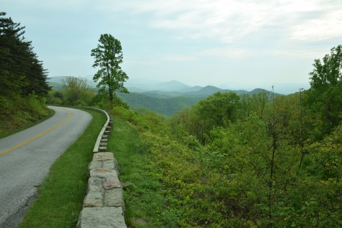 A winding road alongside a stone barrier, with lush green hills and mountains in the background under a cloudy sky.
