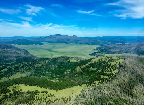 Aerial view of a vast green valley surrounded by mountains under a clear blue sky with scattered clouds.