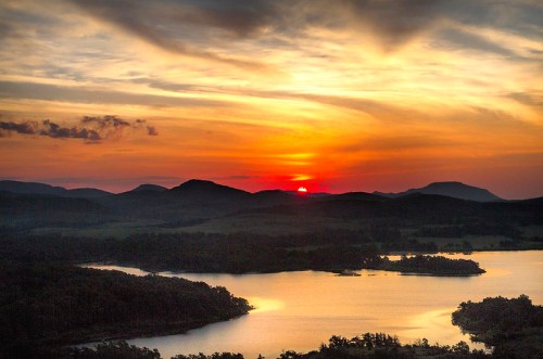 A serene sunset over mountains and a lake, with vibrant orange and yellow hues in the sky.
