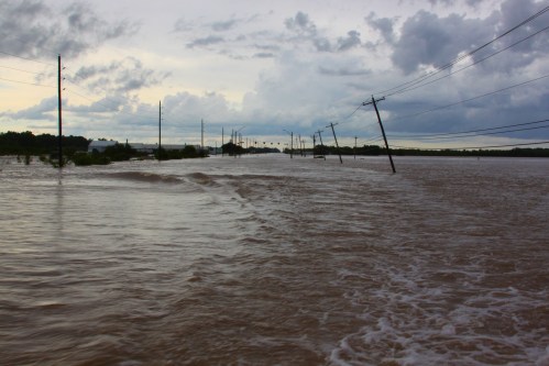 Flooded landscape with submerged roads and leaning power poles under a cloudy sky.