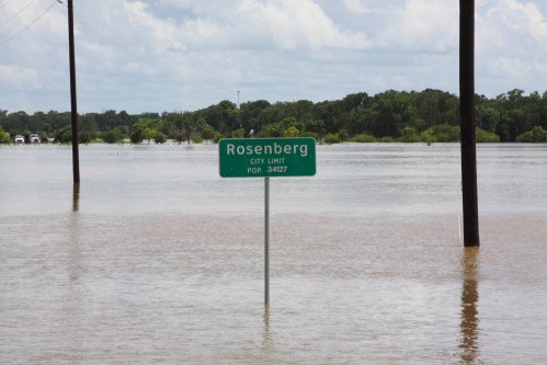 A flooded area with a city limit sign for Rosenberg, indicating a population of 34,127, partially submerged in water.