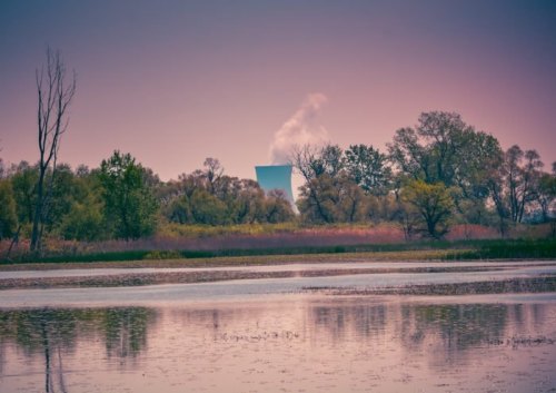 A tranquil landscape with a lake, trees, and a cooling tower emitting steam in the background at dusk.