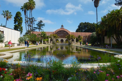A scenic view of a garden with a pond, surrounded by palm trees and colorful flowers, with people walking nearby.