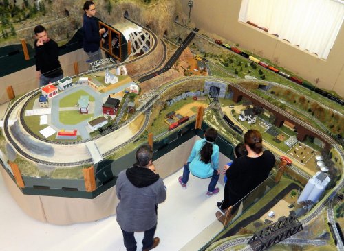 A model train setup with people observing, featuring tracks, buildings, and scenic landscapes.