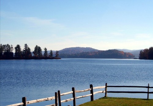 A serene lake view with a wooden fence, surrounded by trees and distant mountains under a clear blue sky.