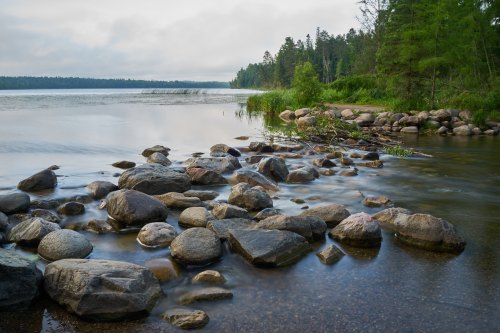 A serene lakeside scene with smooth stones, calm water, and lush greenery along the shore under a cloudy sky.
