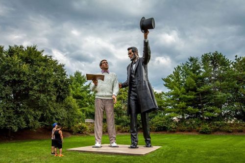 A large statue of Abraham Lincoln stands with a man in a sweater, both against a cloudy sky in a green park.