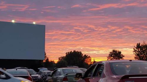 A vibrant sunset with colorful clouds over a drive-in movie theater, surrounded by parked cars.