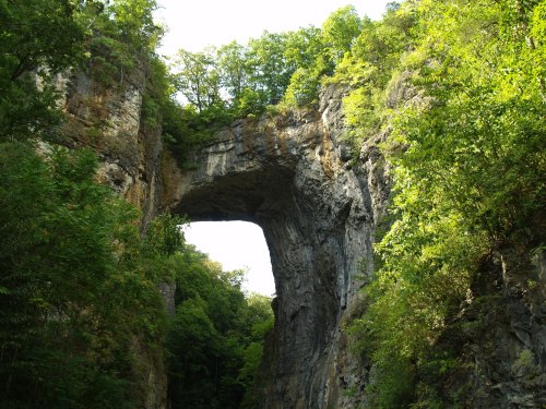 A natural stone arch surrounded by lush green trees and rocky cliffs under a bright sky.