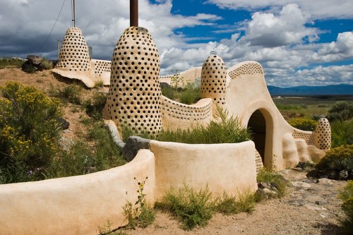 Unique adobe structure with rounded forms and dotted patterns, surrounded by desert vegetation and a cloudy sky.