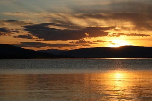 Sunset over a calm lake, with vibrant orange and yellow hues reflecting on the water and silhouetted mountains in the background.