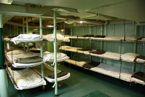 Interior of a ship's bunk room with multiple metal-framed beds, each with a mattress and bedding, arranged in rows.