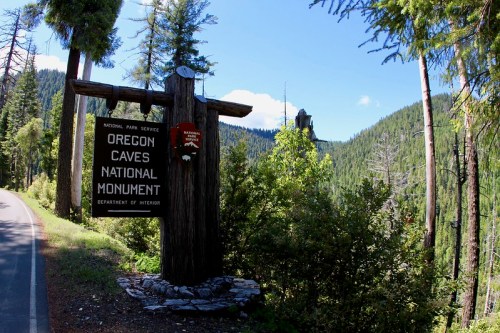 Sign for Oregon Caves National Monument surrounded by lush green trees and mountains under a clear blue sky.