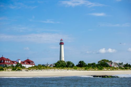 A tall lighthouse with a red top stands near a sandy beach, surrounded by greenery and a clear blue sky.