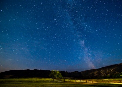A starry night sky with the Milky Way visible above a grassy field and distant mountains, framed by a wooden fence.
