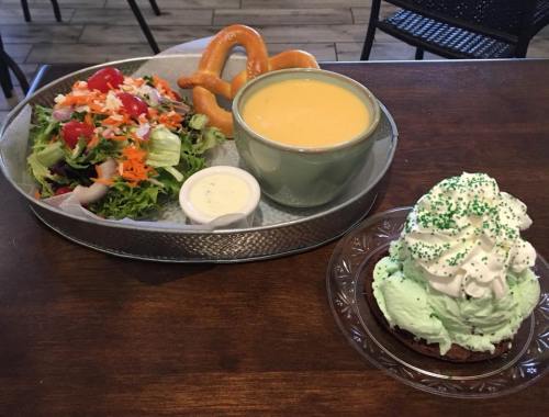A tray with a salad, a bowl of cheese soup, a pretzel, and a dessert with green frosting and whipped cream.
