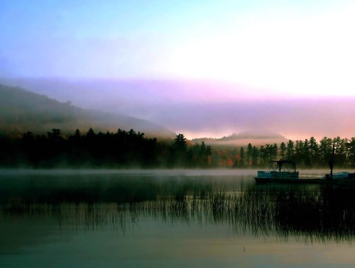 Serene lake at dawn, with mist rising over water and mountains, surrounded by trees and a small boat docked nearby.