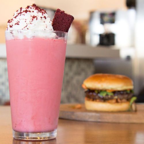 A pink milkshake topped with whipped cream and a red velvet cake piece, with a burger in the background.