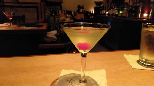 A martini glass with a light green drink and a pink garnish, set on a wooden bar with dim lighting in the background.
