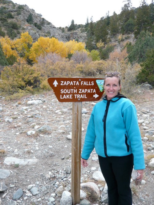 A woman in a blue jacket stands next to a trail sign for Zapata Falls and South Zapata Lake Trail in a mountainous area.