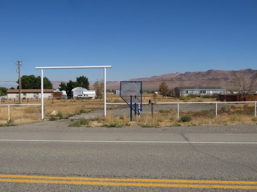 Abandoned lot with a white frame structure, overgrown grass, and distant mountains under a clear blue sky.