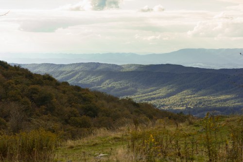 A panoramic view of rolling green hills and mountains under a cloudy sky, showcasing layers of distant peaks.