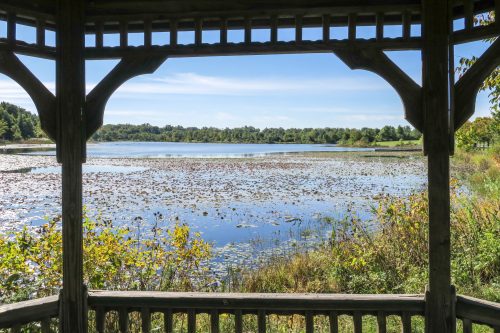 View of a serene lake surrounded by greenery, framed by a wooden gazebo structure.