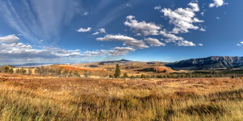 A panoramic view of rolling hills and mountains under a blue sky with scattered clouds, featuring vibrant grasslands.