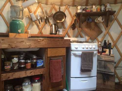 Cozy kitchen in a yurt, featuring a stove, wooden shelves, jars of spices, and a water dispenser.