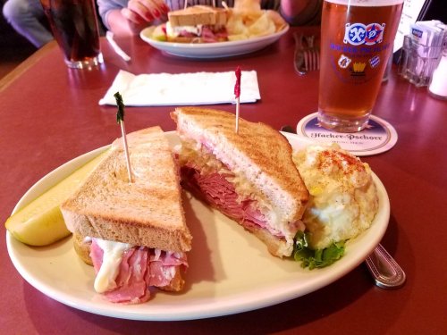 A sandwich with ham and cheese, garnished with toothpicks, served with a side of potato salad and a drink.