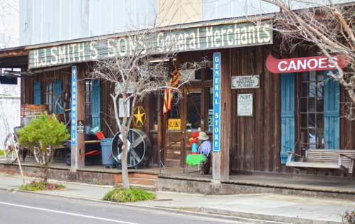 A rustic general store with wooden siding, colorful signs, and a porch, featuring items like canoes and paint.