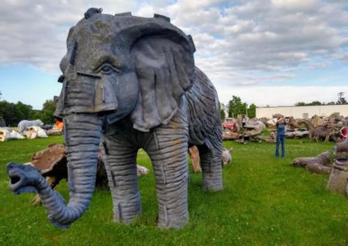 A large, gray elephant sculpture stands in a grassy area, surrounded by various other animal sculptures under a cloudy sky.