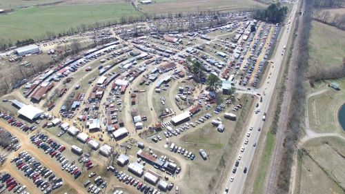 Aerial view of a large outdoor market with numerous stalls, vehicles, and a winding road nearby.