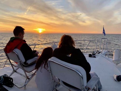 A family sits on a boat, watching a beautiful sunset over the ocean, with calm waters and colorful skies.
